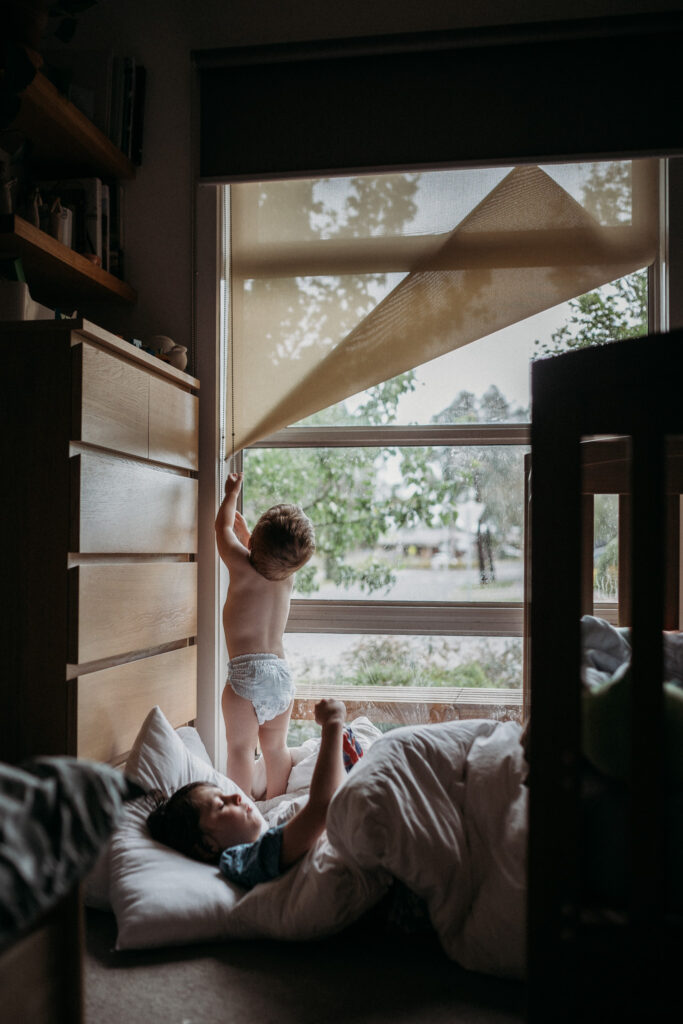 Infant learning to work the roller blinds on window of bedroom while younger brother lies on doona covers on the floor