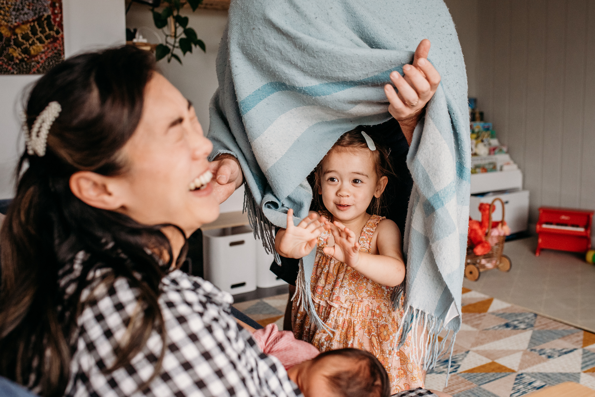 Child hiding under blanket with dad and making mum laugh as she feeds her baby